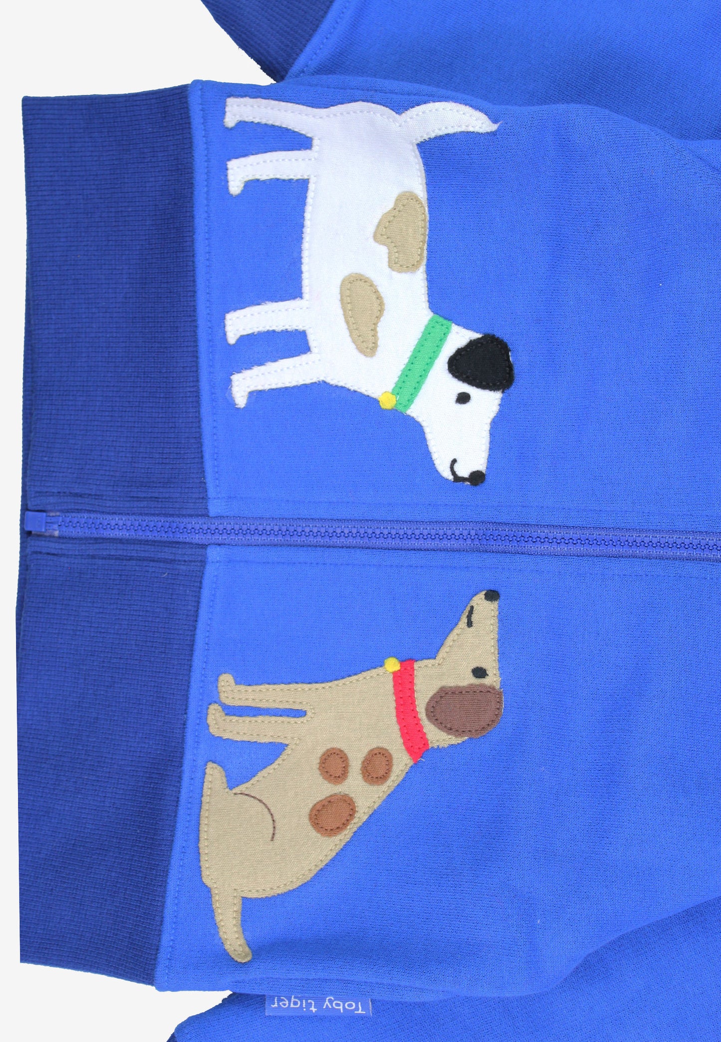 Close-up of a bright blue zippered garment, likely a hoodie or jacket, featuring two embroidered dogs. The top dog is white with tan patches and a green collar, while the bottom dog is tan with brown spots and a red collar.