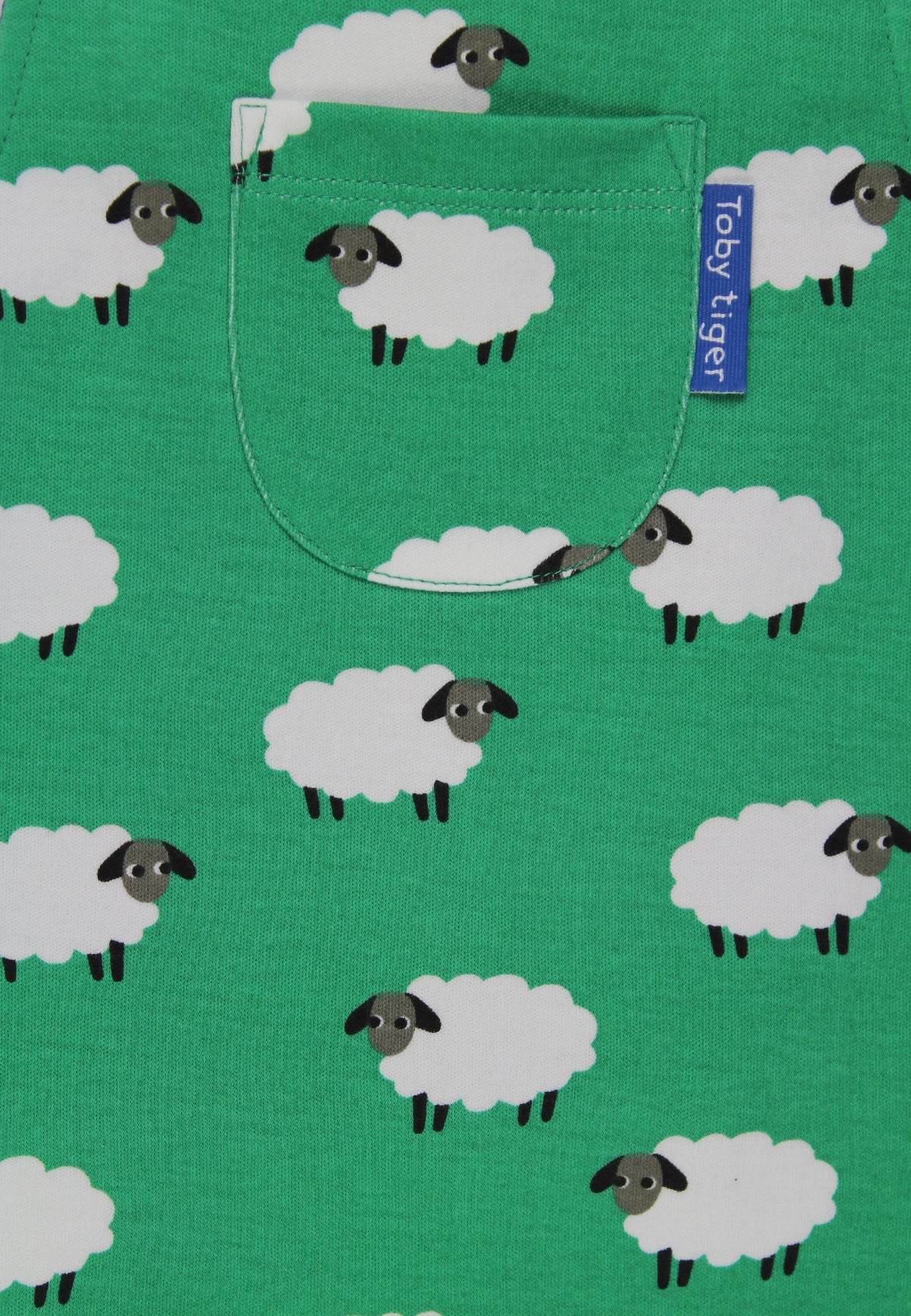 A close-up of a green fabric product, likely a pocket T-shirt, featuring a repeating pattern of white sheep with black faces and legs. A blue fabric tag on the pocket reads 'Toby Tiger'.