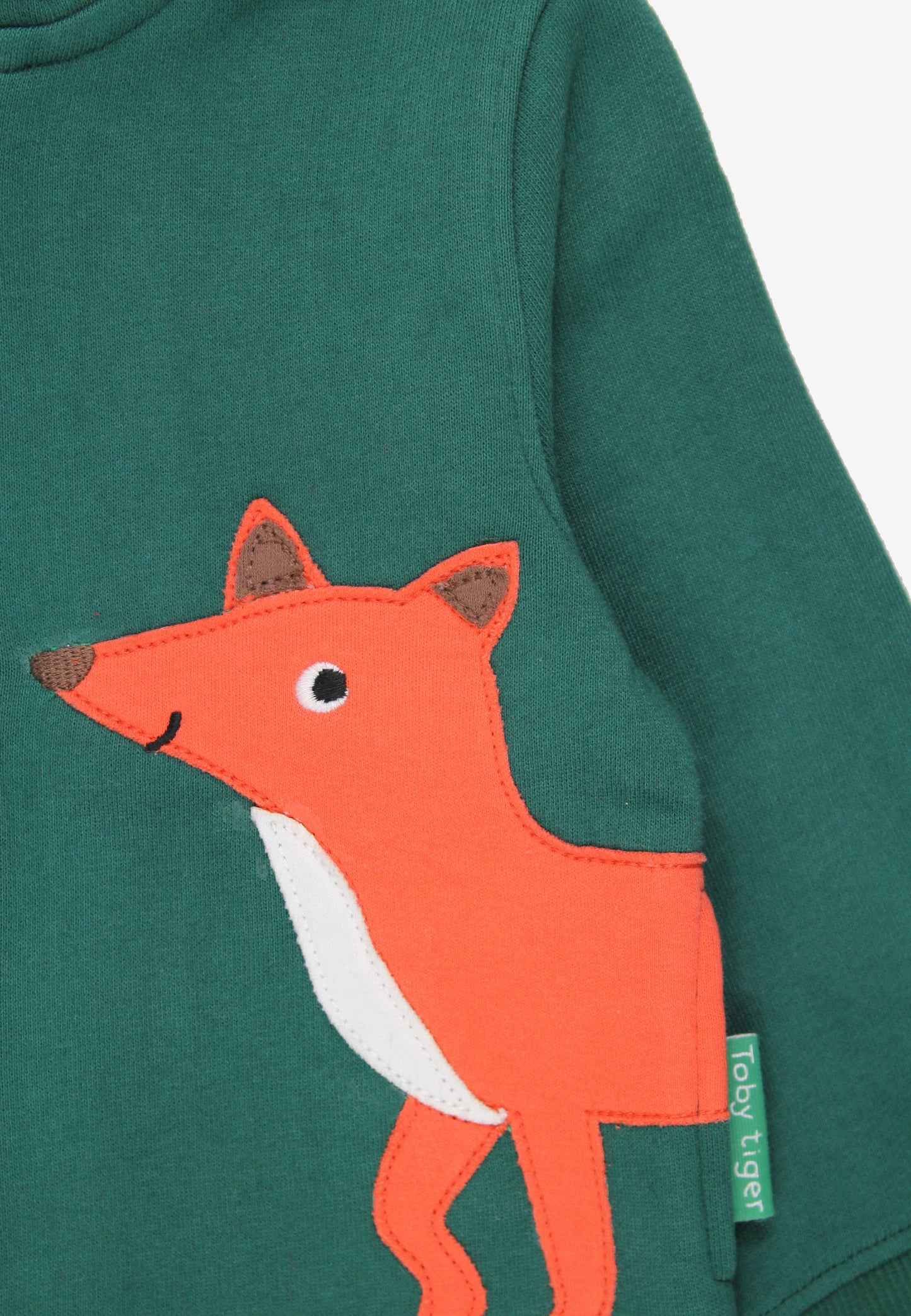 Close-up of a dark green long-sleeve top featuring a large orange fox applique on the front, with a small green fabric tag reading 'Louise Nigra' visible near the sleeve.
