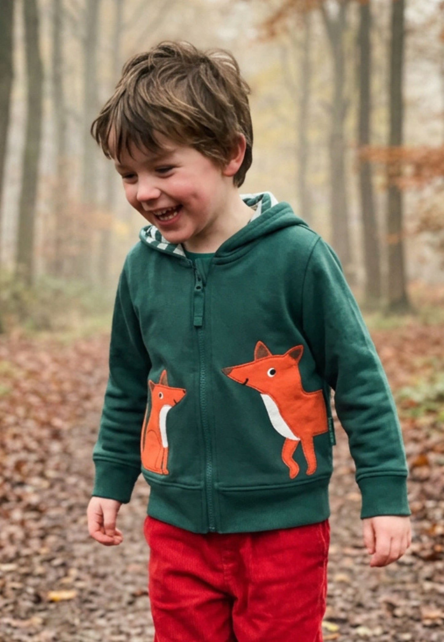 A young boy smiling in an outdoor forest setting, wearing a dark green zip-up hoodie with two orange fox graphics on the front and red pants.