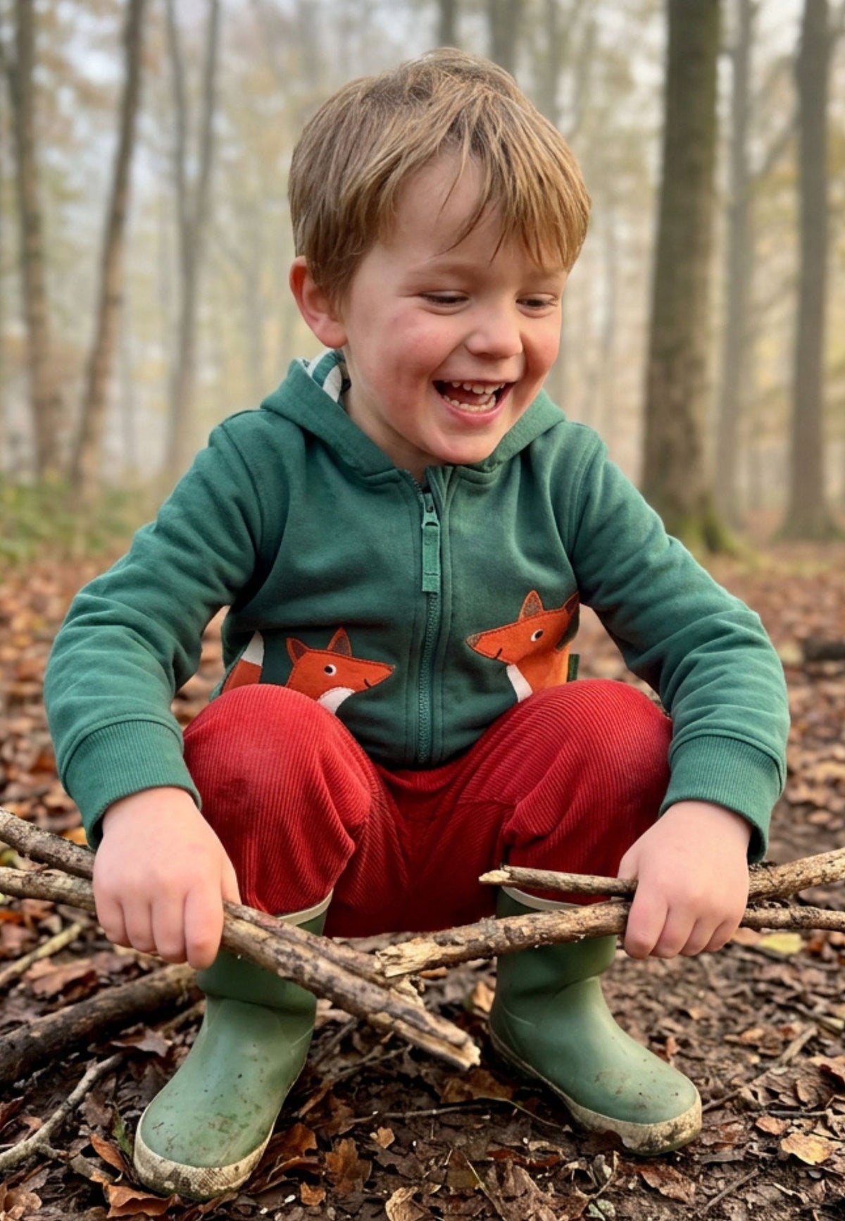 A young boy smiling while crouching outdoors in a wooded setting, wearing a green zip-up hoodie with fox graphics, red pants, and green rain boots.