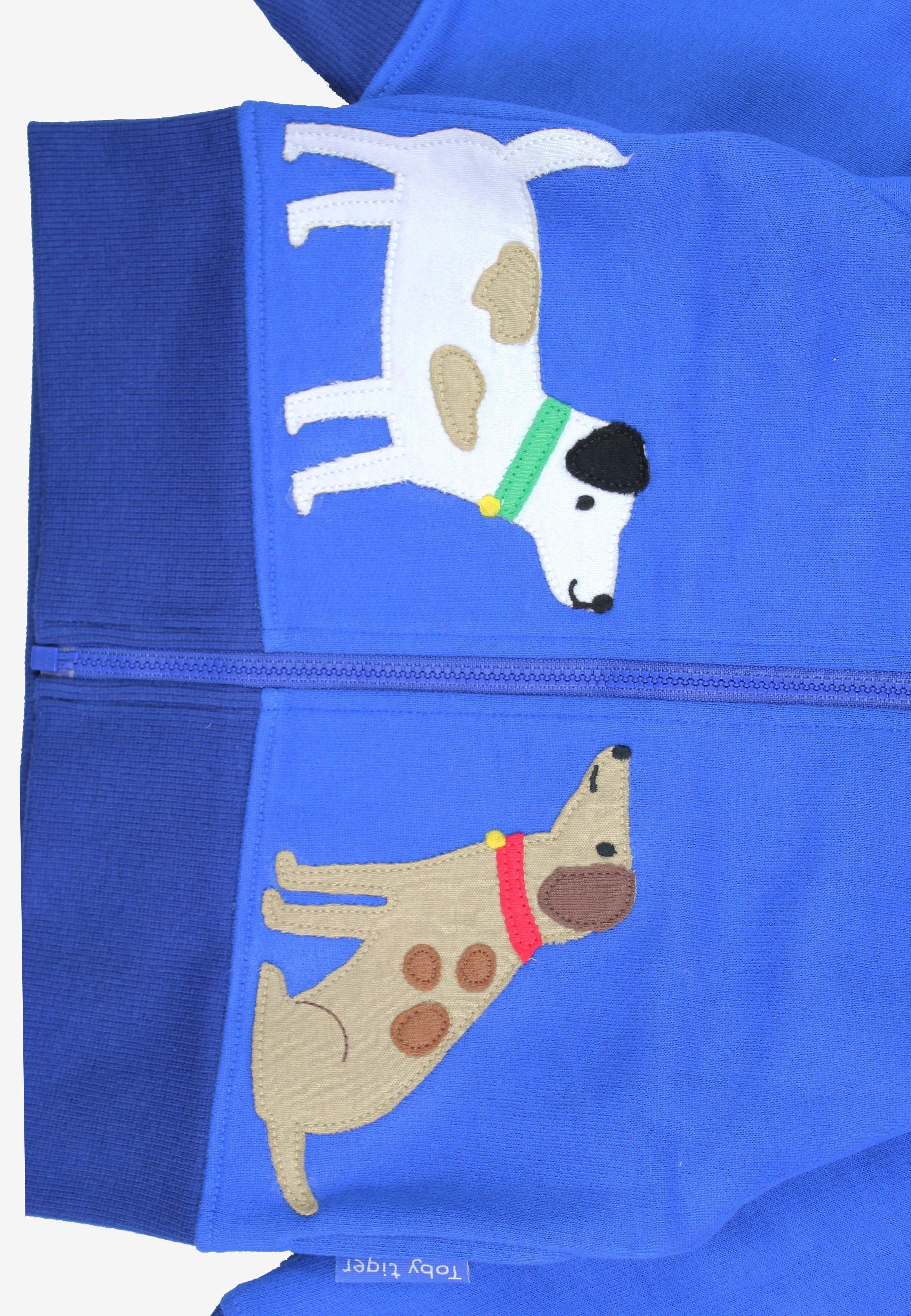 Close-up of a bright blue zippered garment, likely a hoodie or jacket, featuring two embroidered dogs. The top dog is white with tan patches and a green collar, while the bottom dog is tan with brown spots and a red collar.