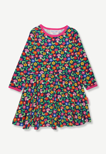 A long-sleeve tiered dress for toddlers or young girls, featuring a dense multicolor floral print on a dark green background with bright pink, red, yellow, and blue flowers, finished with a contrasting pink collar and cuffs.