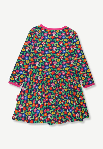 A long-sleeve children's dress with a dense multicolor floral print on a dark green background, featuring bright pink collar and cuff trim, shown flat against a light gray background.