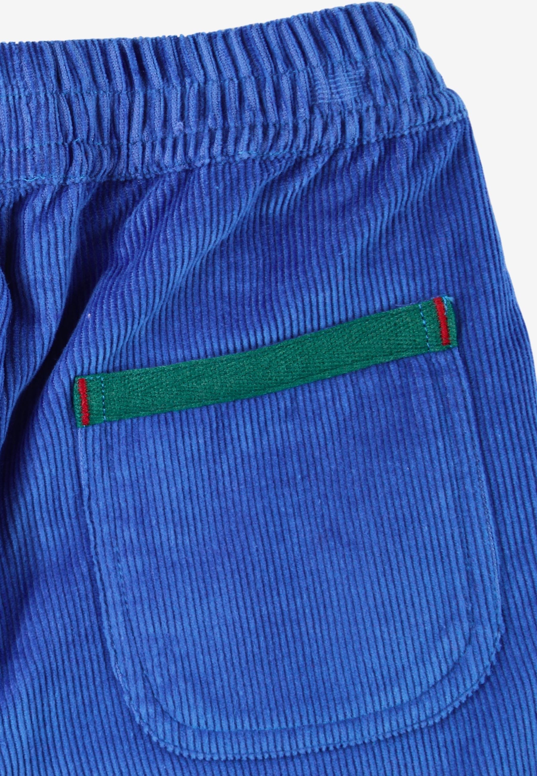 Close-up of blue corduroy trousers showing a back pocket with a green stripe detail bordered in red, on a white background.