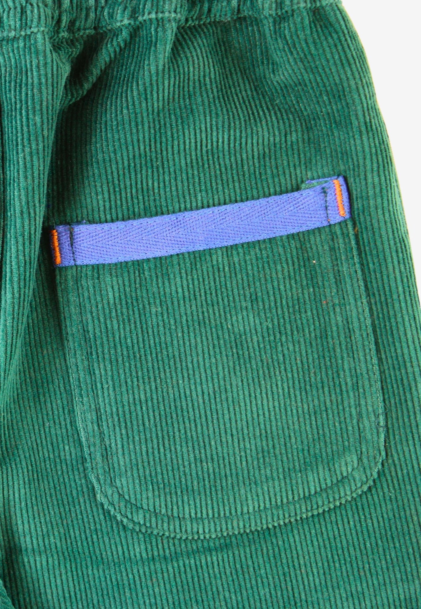 Close-up of a green corduroy garment, likely a jacket or coat, featuring a front patch pocket with a contrasting purple zipper and small orange accent tabs.