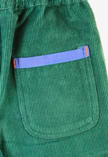 Close-up of a green corduroy garment, likely a jacket or coat, featuring a front patch pocket with a contrasting purple zipper and small orange accent tabs.